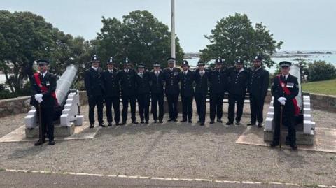 Group of uniformed personnel standing in a line between two large cannons on a raised stone platform, with trees and the sea visible in the background.