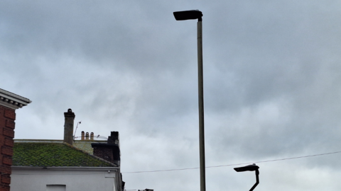 Two street lights on the right. The sky is dark and cloudy. There are houses on the left. 