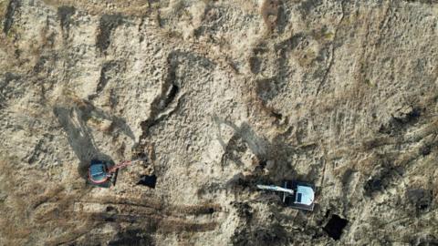 An aerial view looking down on a dry yellow-y brown part of the moor, which has hills and divots. There are also the tracks of two diggers which are visible in the lower half of the image, one has a dark grey roof and orange digger arm while the other is light grey