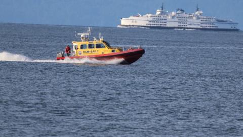 A yellow and red search and rescue boat drives in open waters with a large white vessel in the background