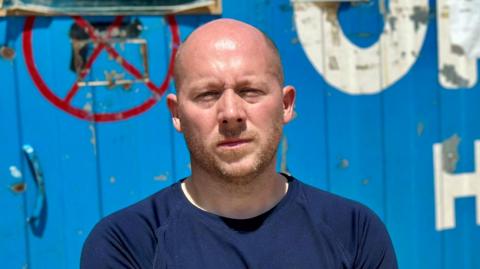 Mr Sears is standing outside a blue shipping container that has write writing on the front. He is wearing a blue top, has some facial hair, and is squinting slightly into the sun. He is not smiling.