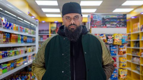 Guz Khan, playing Mobeen, stands in a shop wearing a jacket looking at the camera. He is looking stern and has hat, beard and big jacket in the comedy show.