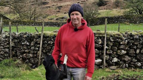 Paddy Deady smiling into the camera. He is standing in a field in front of a stone wall. He is wearing a dark beanie and red hoodie. A black and white dog has jumped up and is standing on its hind legs with front paws on Paddy's waist.
