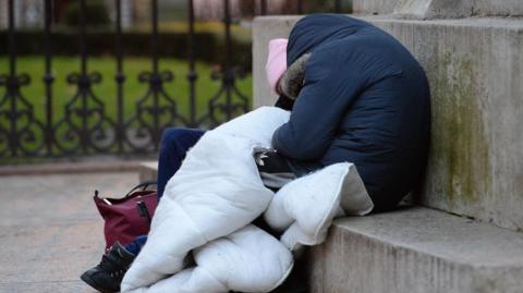 A person sleeping rough slumped against some stone steps. They're wearing a navy blue winter coat and have a white blanket covering their lap.