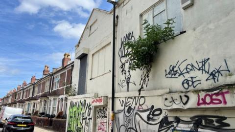 An empty property in St Werburgh's, Bristol. The building is covered in graffiti and there is a row of terraced houses beside it.