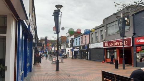 The pedestrianised street is bordered by various shop fronts, including a red shop with a sign that reads: "The Blandford". Red and grey bricks on the road are arranged in a zigzag pattern. Above, large, colourful circular bunting is hanging. There are people coming in and out of the shops.