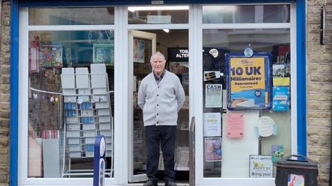 An elderly man in a grey cardigan stands proudly outside the door of a newsagents