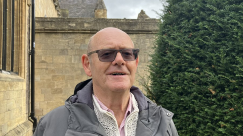 Bald smiling man wearing sunglasses. He is wearing a grey hooded anorak/jacket and cream cardigan with a pink and white striped shirt. He is standing in front of a church and a Yew bush. The sky is cloudy and grey