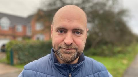 A profile image of Christopher Mallor, a bald man with a short greying beard wearing a blue, padded gilet.