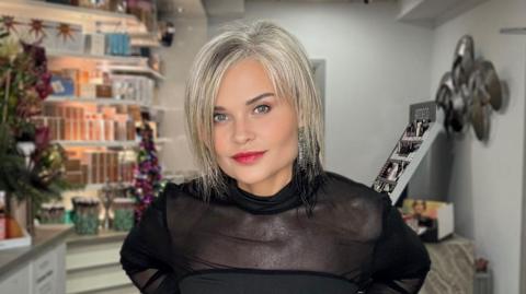 Rosemary has shoulder-length metallic grey hair parted in the middle. She is wearing a black top and behind her is a beauty salon, with a floral arrangement on the counter-top and products on the shelves,