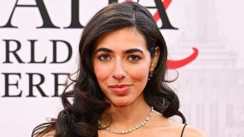 Izzy Escobar wears a black strappy formal dress with black gloves on the red carpet of the Devil Wears Prada 2 premiere. She has long wavy black hair and wears a silver necklace. She stands against a backdrop showing the film title