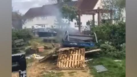 A picture of Springwell Community Hub's memory garden after the fire. Pallets are stacked haphazardly in a pile and smoke is billowing from it.