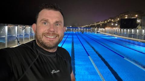 Will Flockton smiling, looking down the camera, with a swimming pool over his shoulder