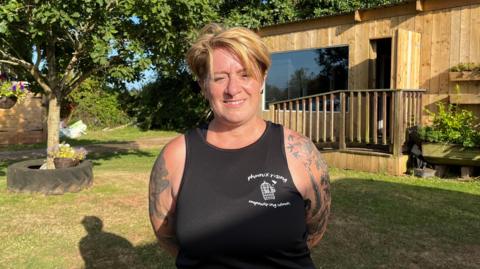 A woman with short blonde hair and a black singlet. She has tattoos on her arms. 