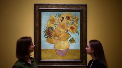 Two women observe Vincent van Gogh's famous painting Sunflowers, displayed in an ornate frame on a yellow wall.