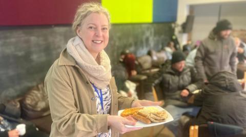 A woman holds a full English breakfast in a large room with people sat at tables and chairs. She is wearing a sand jacket, a black and white top, beige scarf and has long blonde hair tied back. The three men behind her are blurred out of vision and are wearing dark warm clothing including two men in wooly hats. There are leather sofas in the background too positioned against the wall.