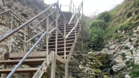 Wooden steps down the side of a cliff that lead to a beach. There are metal handrails down the sides.