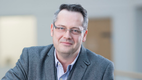 Professor Richard Bowden from the University of Surrey. He is wearing a pale blue shirt and grey suit jacket. He is looking to camera and has greying hair and grey-framed glasses.