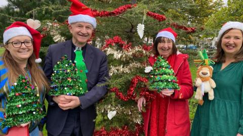 A woman wearing a blue jumper, stood next to a vicar who is standing on one side of a decorated Christmas tree ourdoors. On the other side is a woman wearing a red coat, and a woman wearing a green dress. All four people are wearing red and white Santa hats and are holding mini decorated Christmas trees.