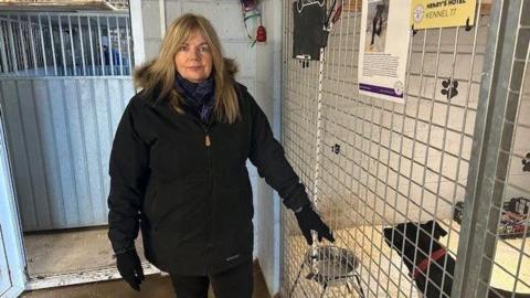 Jane Galliford has long blonde hair and is wearing a black coat with a purple scarf. She is wearing a serious expression on her face. She stands next to a pen with a black dog inside, the dog is looking to the side and it's hard to tell what breed it is.