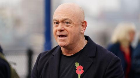 Ross Kemp looking at the camera with his mouth open. He has a shaved head and is wearing a black jacket over a black T-shirt. There is a red poppy pinned to his jacket.