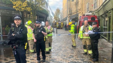 Police office and firefighters standing by blue and white police tape with fire vehicles and a ladder in the background.