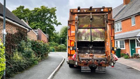 The rear of an orange bin lorry working in a cul-de-sac. Red-brick houses  are on either side.