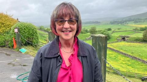 A woman, Jill, wears a pink top and grey jacket. Her hair has pink highlights. She stands in a car park, overlooking a picturesque landscape of fields and trees, looking down a valley.
