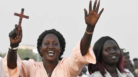 A woman, in a pink top is smiling and holding both hands in the air. Her left hand is open, in a wave, and her right hand is holding a small wooden crucifix
