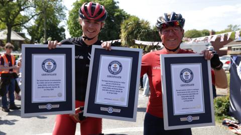 Josh and George Kohler are standing next to each other. They are both wearing red and black cycling wear and helmets. They are holding up three Guinness World Record certificates.