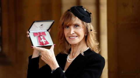 Jackie Bird, who has red hair, smiles at the camera as she shows off her MBE. It is in its case and attached toa ribbon with a bow feature. Jackie is wearing a black jacket and black fascinator. She has a string of pearls around her neck.