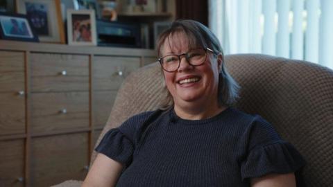 A woman sitting on a chair smiling. She is wearing a navy top with puff sleeves. She has short light brown hair and is wearing glasses.