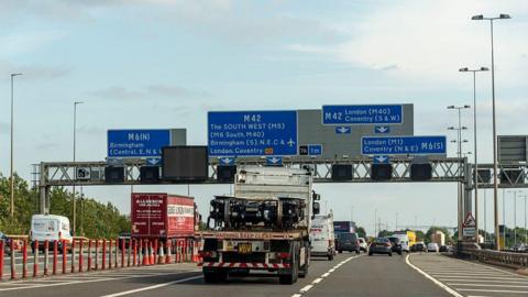 A wide photo of the M6 . On the right there are three lanes merging into two. small lorry with a flat bed is driving behind lines of traffic. A gantry overhead has blue signs reading "M6" and "M42". There is a sign for Birmingham airport on there.