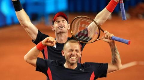 Andy Murray with arms aloft, eyes closed, and a big smile is behind Dan Evans, who celebrates with his racket above his head