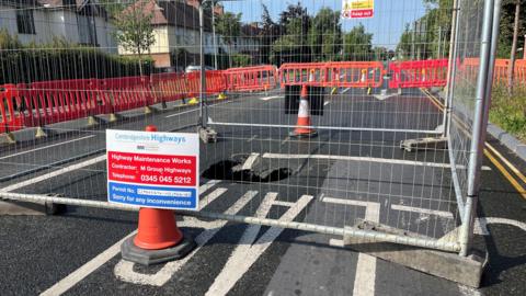 A sinkhole is in the middle of a road which has been closed. Around the sinkhole is a metal fence and cones with signs. Surrounding that are red barriers which have closed the road.