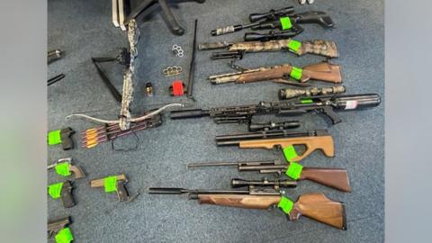 Many weapons laid out on a grey carpet floor. Seven air rifles are in a vertical line to the left of th frame. A crossbow and four arrows are to the right. Just out of frame are what appear to be five handguns. Two knuckledusters are in the middle of the frame. A baton is also among the pile.