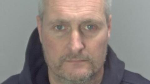 Oliver Arnold has short grey receding hair and a stubbly beard and moustache. He appears on this police custody photo wearing a blue hooded top. The background is grey.