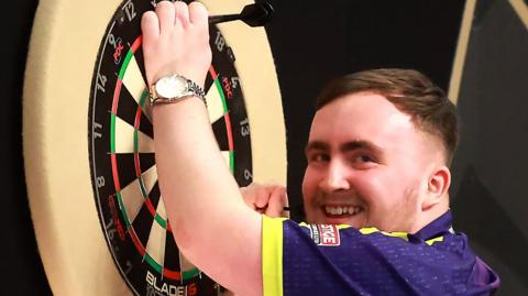 Luke Littler grins as he pulls his darts from the top of a board after winning a game