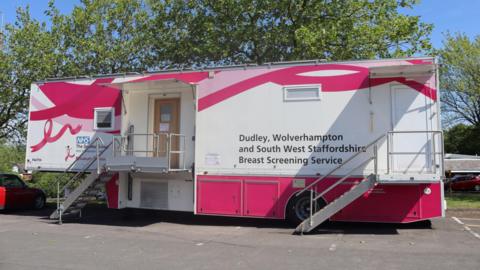 A pink and white breast cancer screening unit in a car park in front of some trees. The unit says 'Dudley, Wolverhampton and South West Staffordshire Breast Screening Service' on the front.