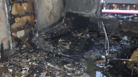 The aftermath of an e-bike fire - a room destroyed by fire with cinders and charred debris littering the floor amid pools of dirty water. 