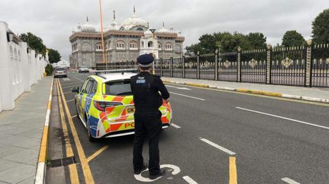 A police officer in black police uniform stands in front of a marked police car. In the back ground is the Sri Guru Nanak Darbar Gurdwara, which is a big grey building with white domes on top. It is  surrounded by a black and gold fence.