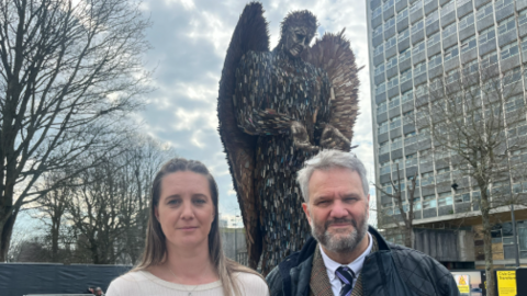 A woman and a man standing side-by-side in front of the Knife Angel.