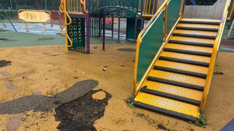 A yellow playground floor has a large chunk ripped out, revealing the earth beneath. There are some yellow steps leading to a slide. In the background you can see a desolate and run down play area. 