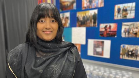 Aishah Ahmed woman is looking right at the camera, she is wearing black clothing with gold sparkles on it. She has dark brown hair and is smiling. She has a nose ring piercing. She is stood in front of a blue wall with photos pinned to it. 