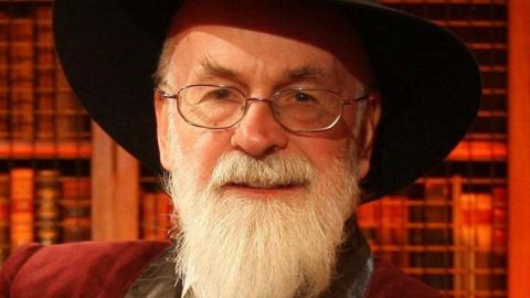 Sir Terry Pratchett is wearing his signature black hat. He has a tidy but long white beard. A bookshelf is stood behind him basked in orange light.