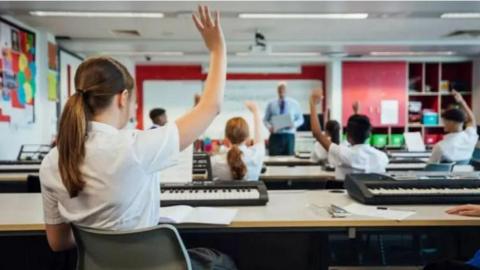 An image of a girl at the back of a classroom with her hand in the air. The image is taken from behind.