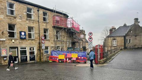 Businesses on Hallstile Bank in Hexham made from sandstone bricks. There is scaffolding, a red telephone box and construction fencing.