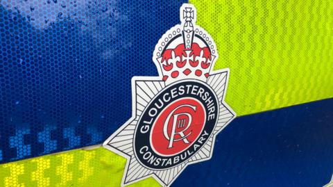The Gloucestershire Constabulary badge on the blue and yellow reflective door of a police car. It is an octagonal badge with a crown on top, and the Charles Royal emblem in the middle.