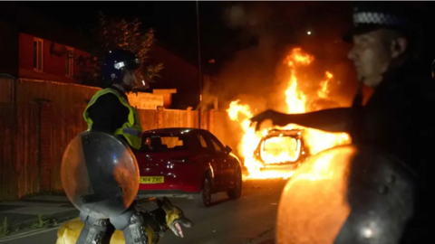 Two police men in riot gear, one with a police dog, standing in front of a burning car.