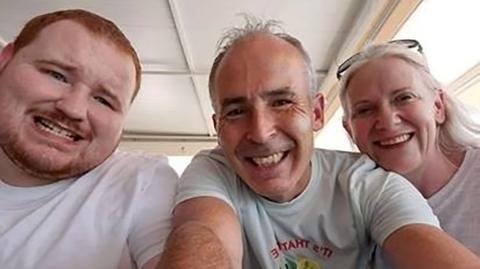 Evan O'Connor and his parents Mark and Louise O'Connor smiling at the camera in a family photo.   Evan has short, ginger hair and a beard.  Mark and Louise both have grey hair.  They are all wearing white t-shirts. 